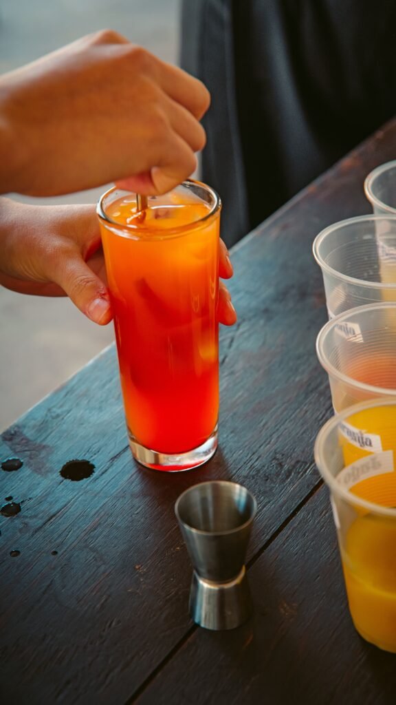 A person stirring a vibrant orange cocktail on a wooden table outdoors within a lively setting.